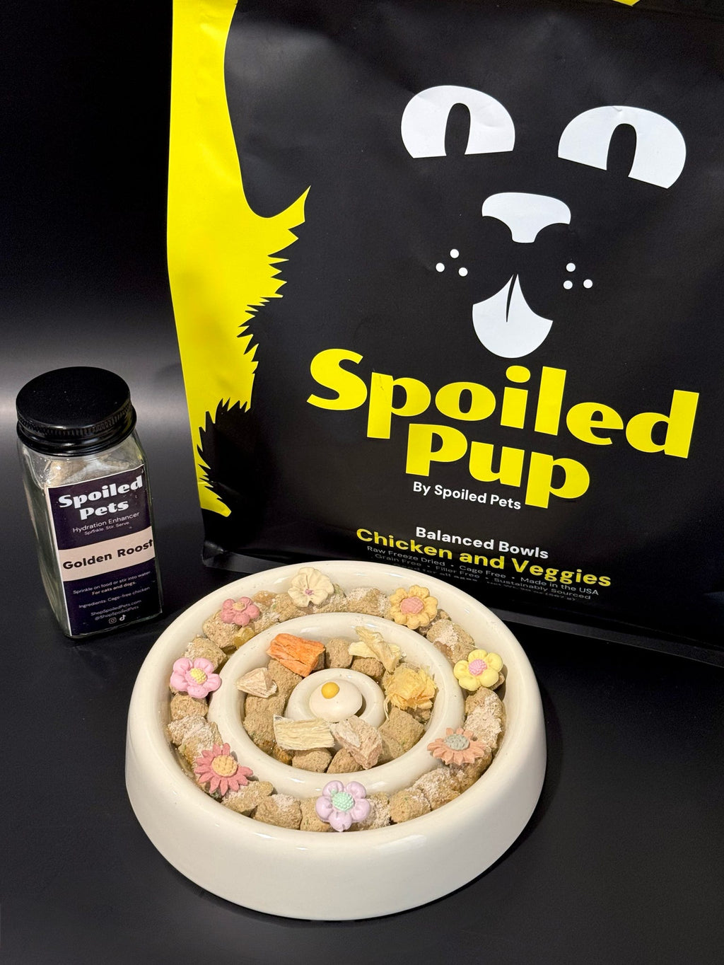 Dog food bowl with treats, a bottle of seasoning, and a 'Spoiled Pup' bag on a black background