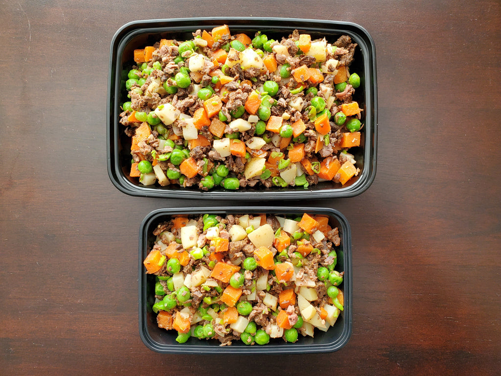 Two black plastic containers filled with a mix of ground meat, carrots, peas, and cauliflower on a wooden surface.