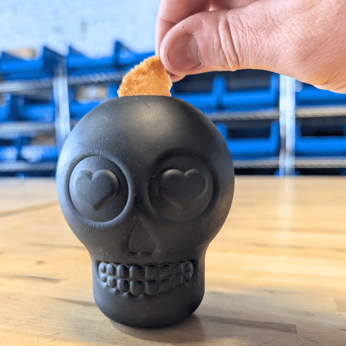 Black skull-shaped object with heart eyes on a wooden surface, held by a hand.