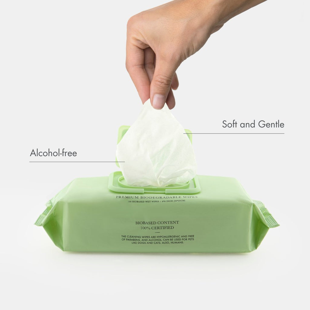 Hand pulling a wet wipe from a green packet of alcohol-free wipes on a white background